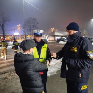 Policjantka i strażnik miejski rozmawiają z kobietą- wręczają jej odblask